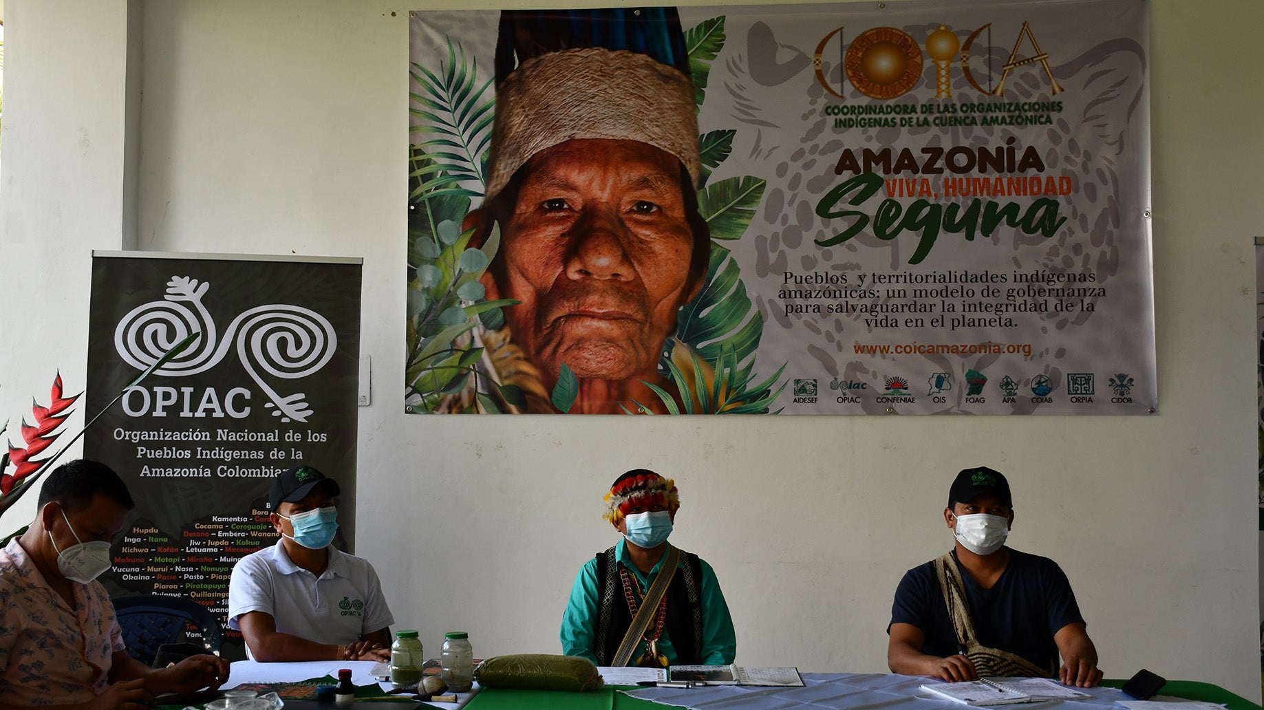 Four individuals with masks sitting at a table with an "OPIAC" sign in the background